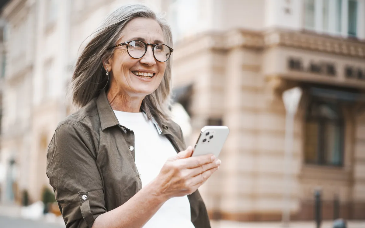 Senhora sorrindo ao ler uma notícia no celular, numa rua da cidade