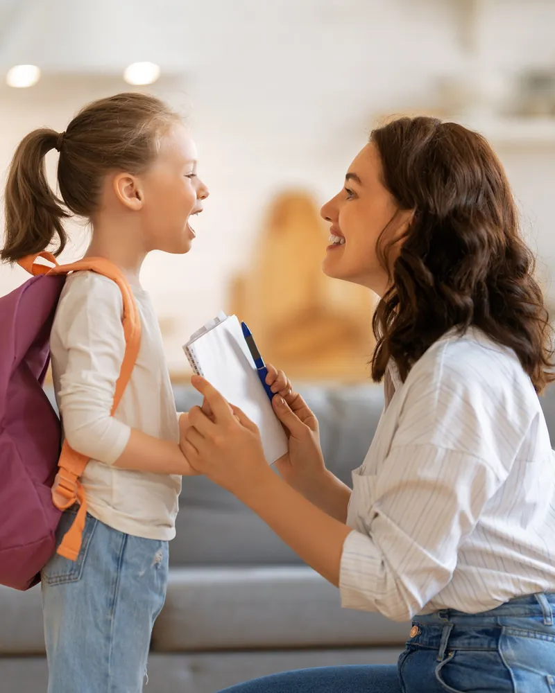 Mãe agachada conversando com a filha de mochila, no dia da volta às aulas