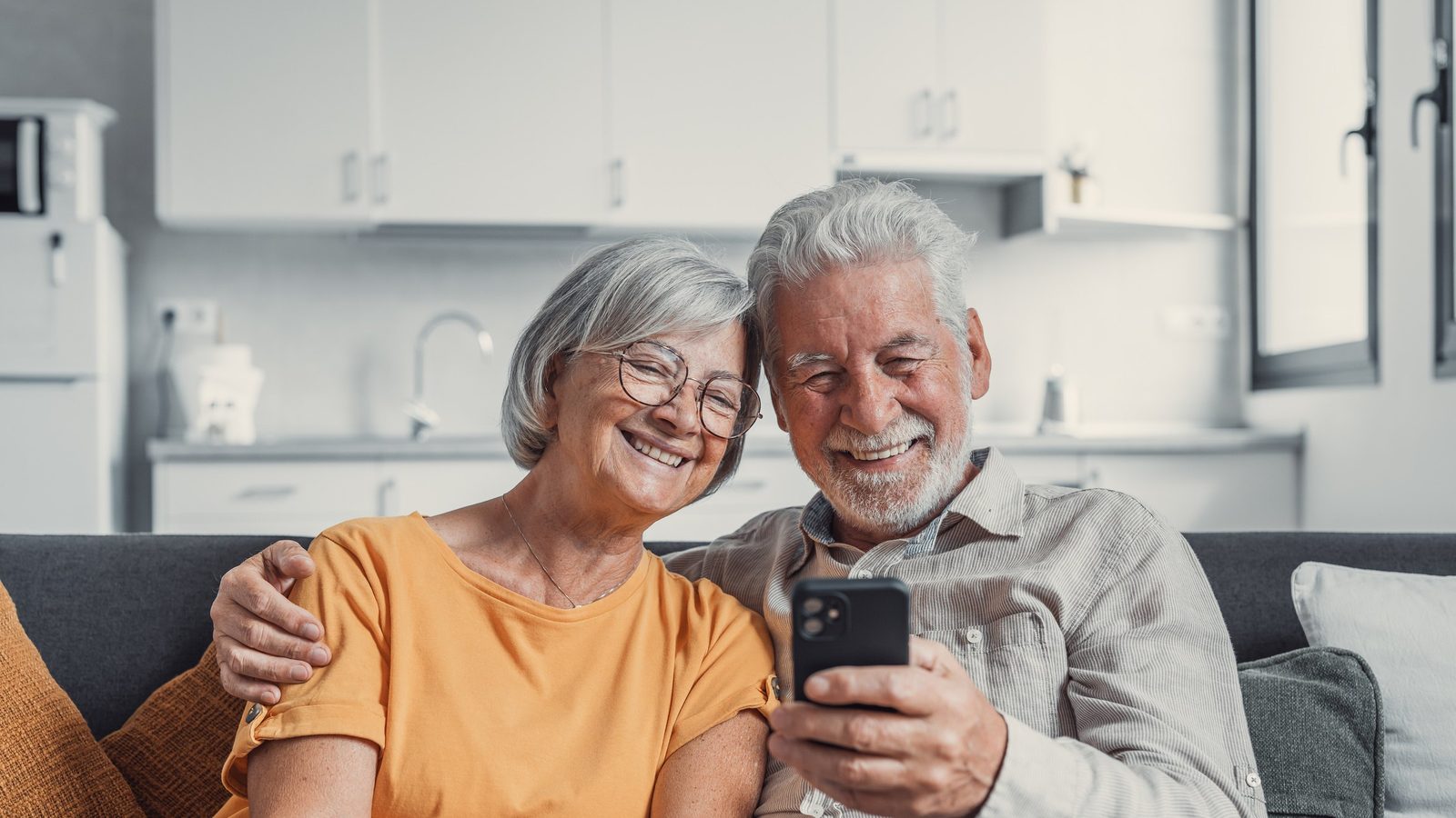 Casal de idosos sorrindo ao ler uma mensagem no celular, no sofá de casa